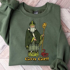 Peut inclure: Sweat-shirt vert avec une image de dessin animé d'un lutin tenant un bâton avec un trèfle au sommet. Le texte "Go'n Git!!" est imprimé sous l'image.