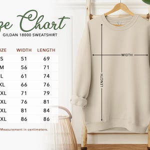 May include: A beige Gildan 18000 sweatshirt displayed on a wooden hanger. The image includes a size chart with measurements in centimeters, detailing width and length for sizes S through 5XL. The sweatshirt is shown with length and width indicators.