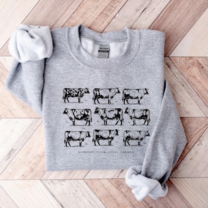 Op de afbeelding: Een lichtgrijs sweatshirt met een zwart-wit illustratie van negen koeien. De tekst "SUPPORT YOUR LOCAL FARMER" is onder de koeiengrafiek gedrukt. Het sweatshirt is op een houten oppervlak gevouwen.