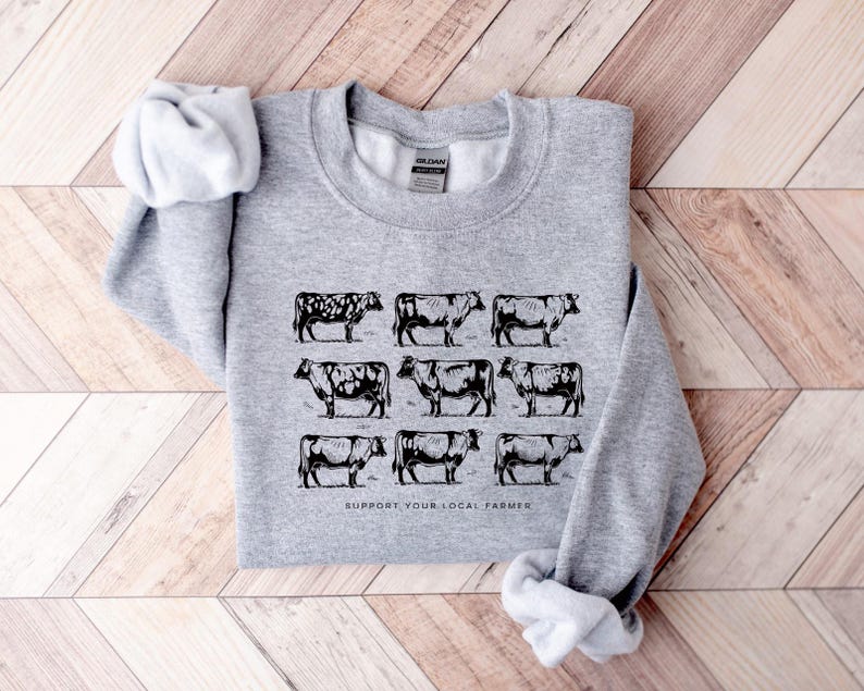 Op de afbeelding: Een lichtgrijs sweatshirt met een zwart-wit illustratie van negen koeien. De tekst "SUPPORT YOUR LOCAL FARMER" is onder de koeiengrafiek gedrukt. Het sweatshirt is op een houten oppervlak gevouwen.