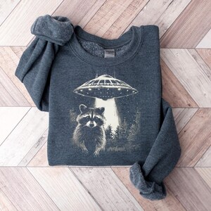May include: A dark gray sweatshirt featuring a graphic of a raccoon standing in front of a forest with a UFO shining a light down on it. The sweatshirt is made of a soft material and has a crew neck.