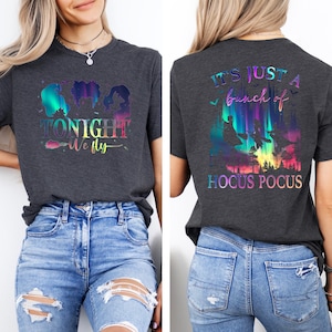 May include: Dark gray t-shirt with a colorful graphic featuring silhouettes of three witches and the text "TONIGHT WE FLY" on the front. The back of the shirt has the text "IT'S JUST A bunch of HOCUS POCUS" with a colorful witchy scene.
