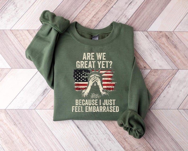 Puede incluir: Sudadera verde con un gr&aacute;fico de la Estatua de la Libertad con los ojos tapados y el texto "Are we great yet? Because I just feel embarrassed."