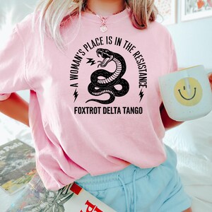 May include: A pink t-shirt with a black snake graphic and the text "A woman's place is in the resistance" and "Fox Trot Delta Tango".