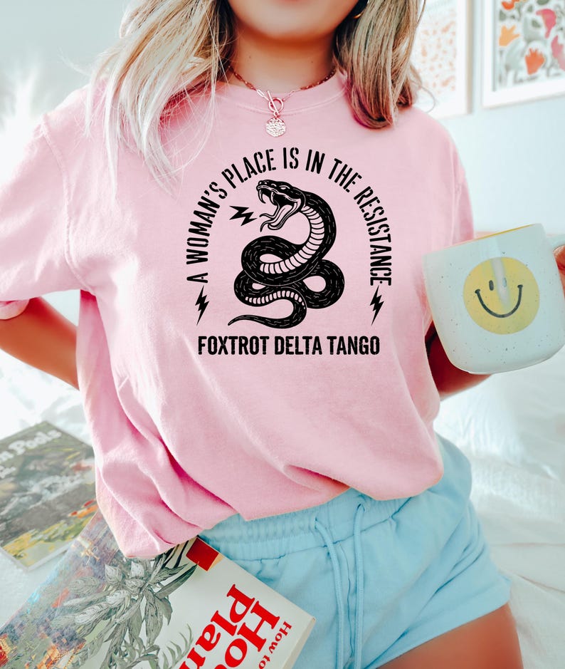 May include: A pink t-shirt with a black snake graphic and the text "A woman's place is in the resistance" and "Fox Trot Delta Tango".