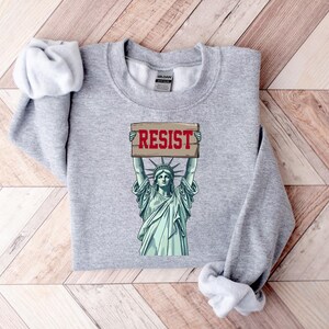 Op de afbeelding: Grijs sweatshirt met een afbeelding van het Vrijheidsbeeld dat een bord vasthoudt met de tekst "RESIST".