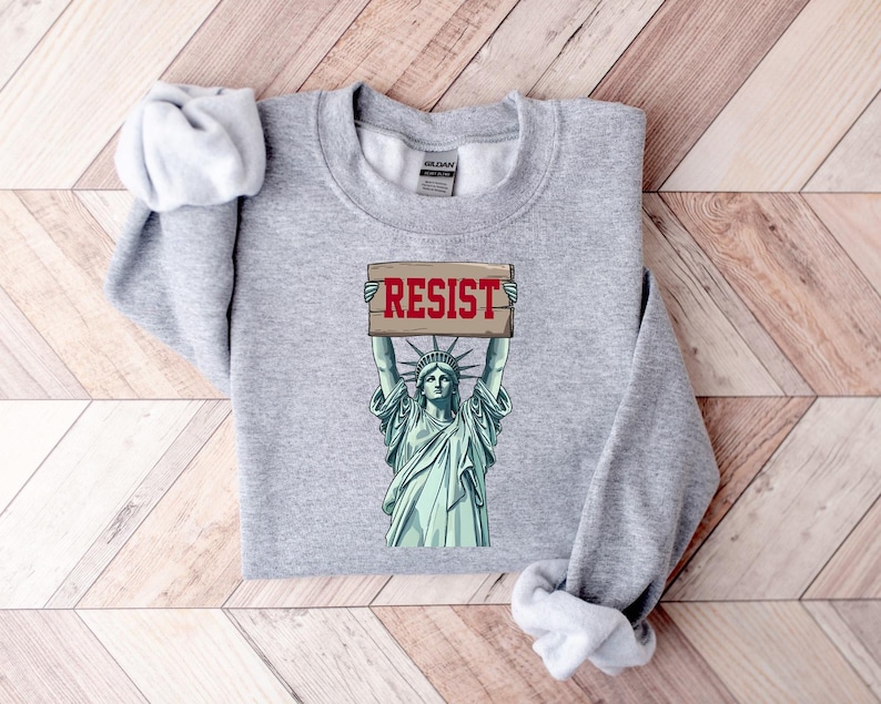 Op de afbeelding: Grijs sweatshirt met een afbeelding van het Vrijheidsbeeld dat een bord vasthoudt met de tekst "RESIST".