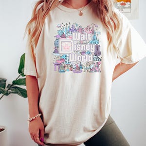 May include: A woman wearing a light beige t-shirt with a graphic print of Walt Disney World landmarks and the text "Walt Disney World". She is also wearing dark green biker shorts.