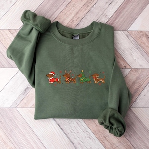 May include: An olive green sweatshirt featuring an embroidered design of four dachshunds dressed in Christmas attire. The dogs are wearing a Santa suit, reindeer antlers, a Christmas tree, and a gingerbread outfit. The sweatshirt has long sleeves.