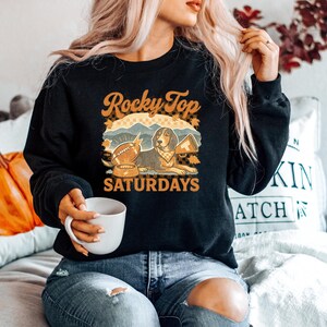 May include: Black sweatshirt with a graphic featuring a dog, football, and the text "Rocky Top Saturdays." The design includes fall leaves and a mountain scene. The sweatshirt is a crew neck style.