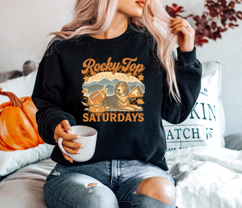 May include: Black sweatshirt with a graphic featuring a dog, football, and the text "Rocky Top Saturdays." The design includes fall leaves and a mountain scene. The sweatshirt is a crew neck style.