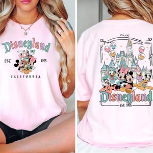 Puede incluir: Camiseta rosa con un gr&aacute;fico de Mickey Mouse y sus amigos frente a Disneyland. La parte delantera de la camiseta dice "Disneyland Est. 1955 California". La parte trasera de la camiseta tiene un gr&aacute;fico de Mickey Mouse y sus amigos frente al Castillo de la Bella Durmiente con el texto "Disneyland Est. 1955".