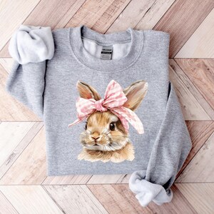 May include: Gray sweatshirt with a brown bunny wearing a pink and white bow. The bunny is looking to the right.