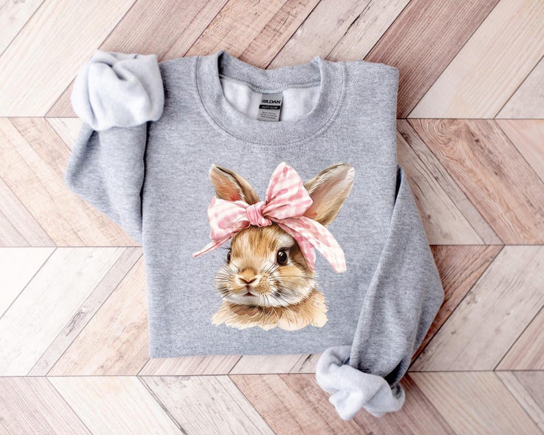 May include: Gray sweatshirt with a brown bunny wearing a pink and white bow. The bunny is looking to the right.