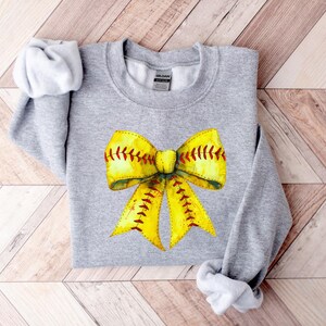 K&ouml;nnte beinhalten: Ein graues Sweatshirt mit einem gelben und roten Softball-Schleifendesign. Die Schleife ist mit einem roten Band gebunden und hat wei&szlig;e N&auml;hte.