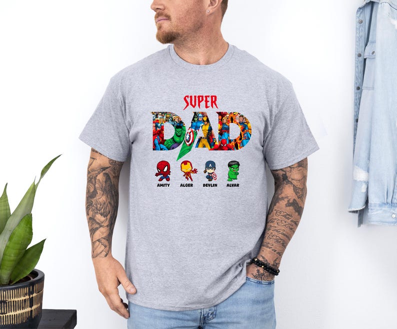 May include: A light grey t-shirt featuring the text "SUPER DAD" in red, with a collage of superhero characters forming the letters. Below, four cartoon superhero figures are labelled Amity, Alger, Devlin, and Alvar. The shirt is made of a soft material.