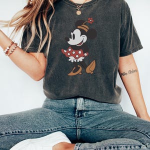 May include: Dark gray t-shirt featuring a graphic of Minnie Mouse in a red polka dot dress, brown shoes, and a hat with a flower. The shirt is paired with blue jeans and white sneakers, creating a casual, everyday look.