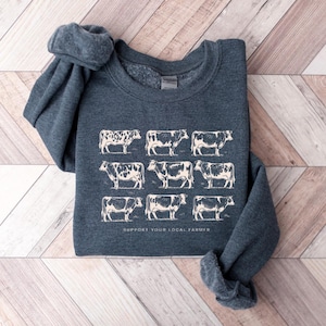 Op de afbeelding: Een donkerblauwe sweatshirt met een cr&egrave;mekleurige illustratie van negen koeien. De tekst "SUPPORT YOUR LOCAL FARMER" staat onder de koeien gedrukt. De sweatshirt ligt op een houten oppervlak.