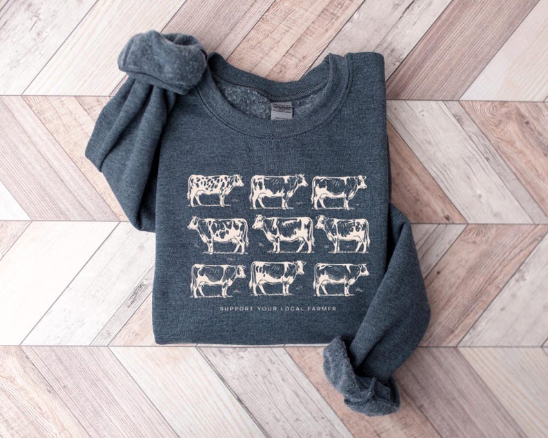 Op de afbeelding: Een donkerblauwe sweatshirt met een cr&egrave;mekleurige illustratie van negen koeien. De tekst "SUPPORT YOUR LOCAL FARMER" staat onder de koeien gedrukt. De sweatshirt ligt op een houten oppervlak.