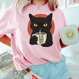 May include: Pink t-shirt with a black cat illustration holding a coffee mug that says "Is he dead yet?"