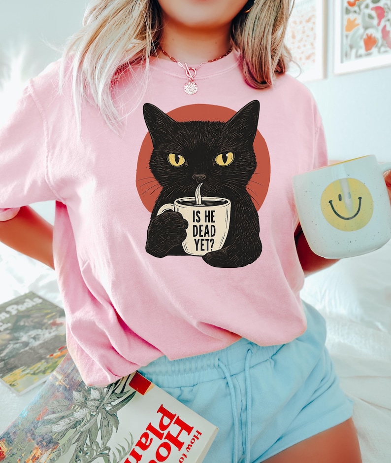May include: Pink t-shirt with a black cat illustration holding a coffee mug that says "Is he dead yet?"