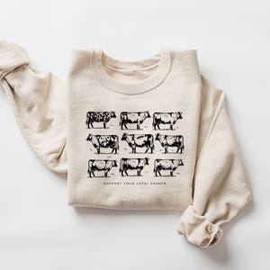Op de afbeelding: Cr&egrave;mekleurige sweatshirt met een zwarte afbeelding van negen koeien in een rasterpatroon. De tekst "SUPPORT YOUR LOCAL FARMER" staat onder de koeien gedrukt. De sweatshirt heeft een ronde hals en lange mouwen.
