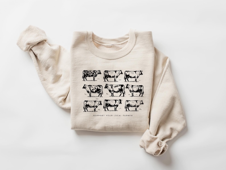 Op de afbeelding: Cr&egrave;mekleurige sweatshirt met een zwarte afbeelding van negen koeien in een rasterpatroon. De tekst "SUPPORT YOUR LOCAL FARMER" staat onder de koeien gedrukt. De sweatshirt heeft een ronde hals en lange mouwen.