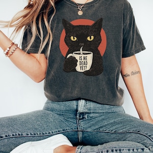 May include: A gray t-shirt with a black cat illustration holding a coffee mug that says "Is he dead yet?" The cat is in front of a red circle.