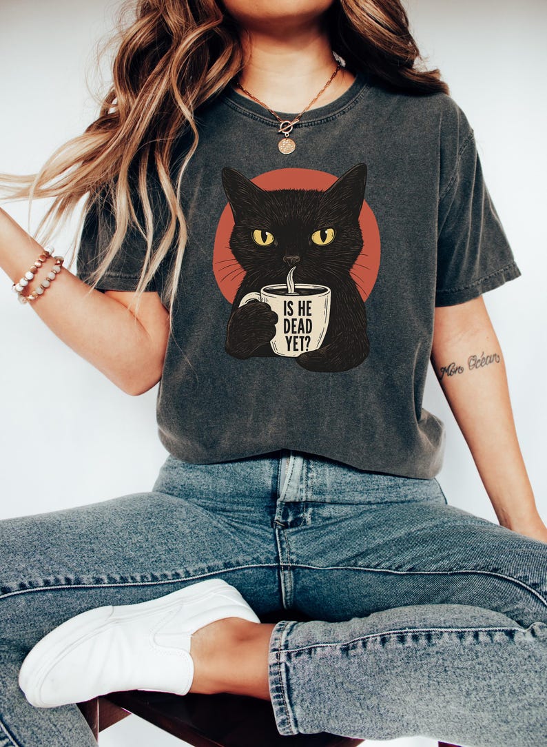 May include: A gray t-shirt with a black cat illustration holding a coffee mug that says "Is he dead yet?" The cat is in front of a red circle.