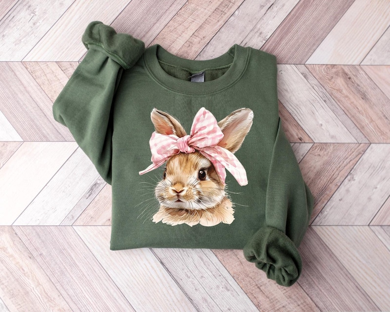 May include: A green sweatshirt with a brown bunny wearing a pink and white polka dot bow. The bunny is looking at the camera.