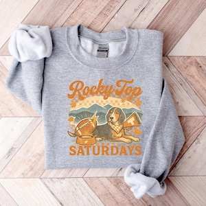 May include: A heather gray sweatshirt with the text "Rocky Top Saturdays" in orange and white. The design features a football, a dog with a megaphone, and mountain scenery.