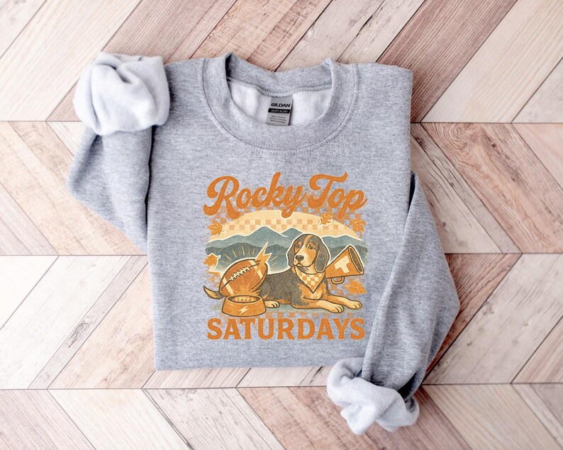May include: A heather gray sweatshirt with the text "Rocky Top Saturdays" in orange and white. The design features a football, a dog with a megaphone, and mountain scenery.