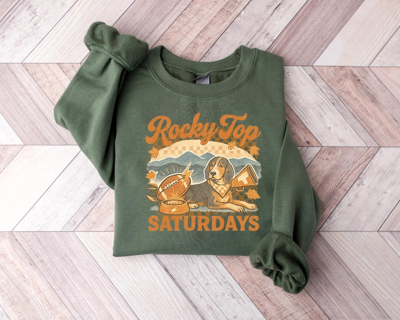 May include: Green sweatshirt with the text "Rocky Top Saturdays" in orange. The graphic features a dog, football, and megaphone. The sweatshirt is folded on a wooden surface.