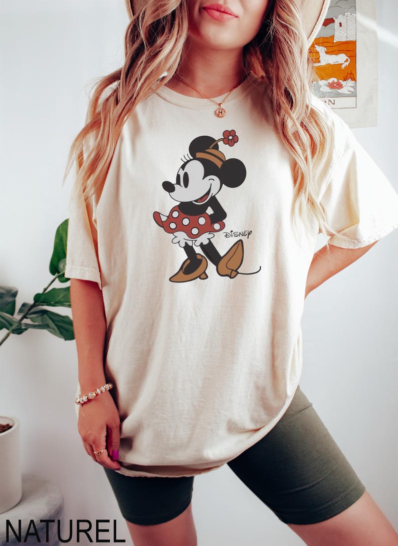 May include: Cream-colored t-shirt featuring a cartoon Minnie Mouse design. Minnie is depicted in a red polka-dot skirt, brown shoes, and a matching hat. The word "Disney" is printed below the character. The word "NATUREL" is printed at the bottom.