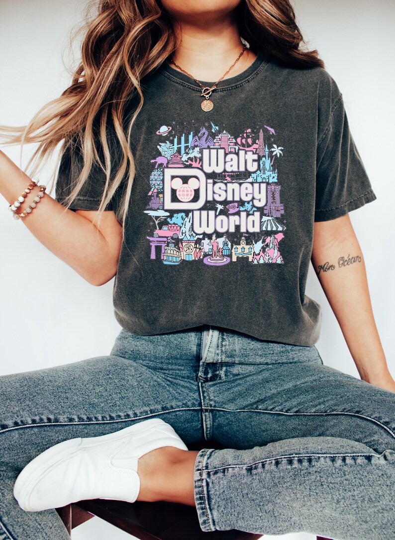May include: A dark gray t-shirt with a graphic print of the Walt Disney World logo surrounded by colorful illustrations of various Disney attractions. The text "Walt Disney World" is printed in pink and blue.