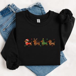 May include: Black sweatshirt featuring an embroidered design of four dachshunds dressed in Christmas attire. The dogs are wearing a Santa hat, reindeer antlers, a Christmas tree, and a festive scarf. The sweatshirt is folded on top of blue jeans.