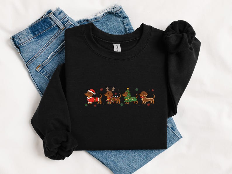 May include: Black sweatshirt featuring an embroidered design of four dachshunds dressed in Christmas attire. The dogs are wearing a Santa hat, reindeer antlers, a Christmas tree, and a festive scarf. The sweatshirt is folded on top of blue jeans.