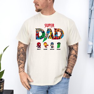 May include: A cream-coloured t-shirt featuring the text "SUPER DAD" in red, with the word "DAD" filled with colourful comic book characters. Below the text are four cartoon characters named Amity, Alger, Devlin, and Alvar.