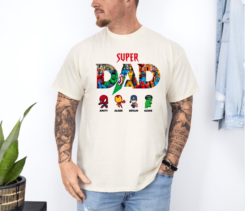 May include: A cream-coloured t-shirt featuring the text "SUPER DAD" in red, with the word "DAD" filled with colourful comic book characters. Below the text are four cartoon characters named Amity, Alger, Devlin, and Alvar.