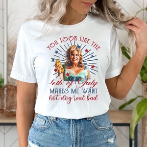 May include: White t-shirt with a red, white, and blue graphic of a woman holding a hot dog. The text on the shirt reads "You look like the 4th of July makes me want hot dog real bad."