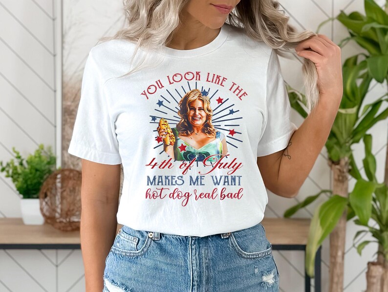 May include: White t-shirt with a red, white, and blue graphic of a woman holding a hot dog. The text on the shirt reads "You look like the 4th of July makes me want hot dog real bad."