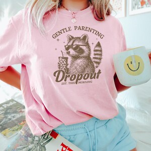 May include: Pink t-shirt featuring a graphic of a raccoon wearing sunglasses and holding a coffee cup. The text on the shirt reads "Gentle Parenting Dropout Est. This Morning".