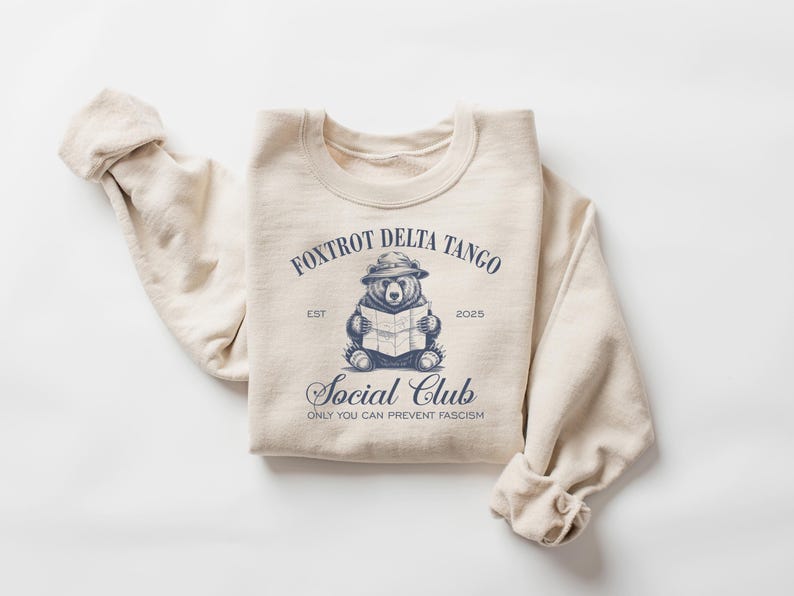Puede incluir: Una sudadera beige con un gr&aacute;fico azul de un oso con un sombrero y sosteniendo un mapa. El gr&aacute;fico incluye el texto "Foxtrot Delta Tango Social Club Est 2023" y "Only You Can Prevent Fascism".
