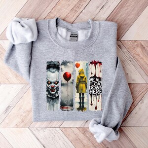 May include: A light gray sweatshirt with a graphic design featuring a scary clown, a person in a yellow raincoat, and the text "WE ALL FLOAT DOWN HERE." The design includes red balloons and dripping blood.