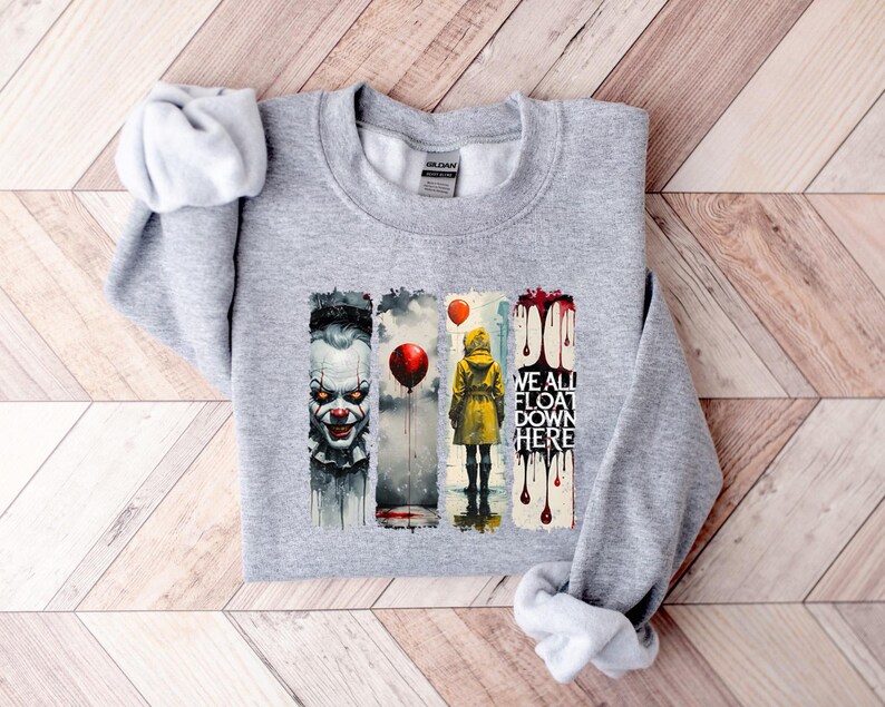 May include: A light gray sweatshirt with a graphic design featuring a scary clown, a person in a yellow raincoat, and the text "WE ALL FLOAT DOWN HERE." The design includes red balloons and dripping blood.