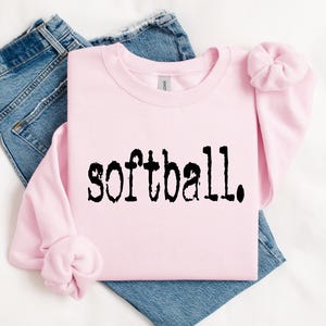 K&ouml;nnte beinhalten: Ein rosa Sweatshirt mit dem Wort "Softball" in Schwarz auf der Vorderseite. Das Sweatshirt ist gefaltet und liegt auf einer wei&szlig;en Oberfl&auml;che mit einer blauen Jeans.
