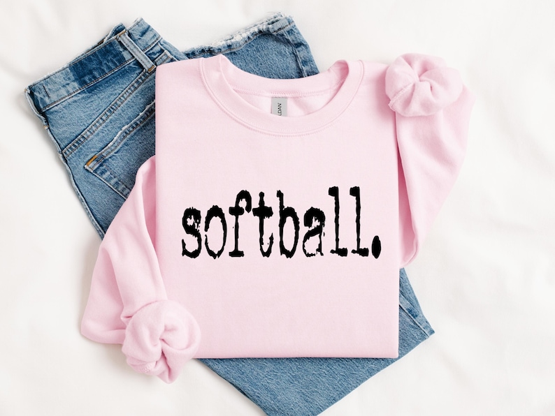 K&ouml;nnte beinhalten: Ein rosa Sweatshirt mit dem Wort "Softball" in Schwarz auf der Vorderseite. Das Sweatshirt ist gefaltet und liegt auf einer wei&szlig;en Oberfl&auml;che mit einer blauen Jeans.