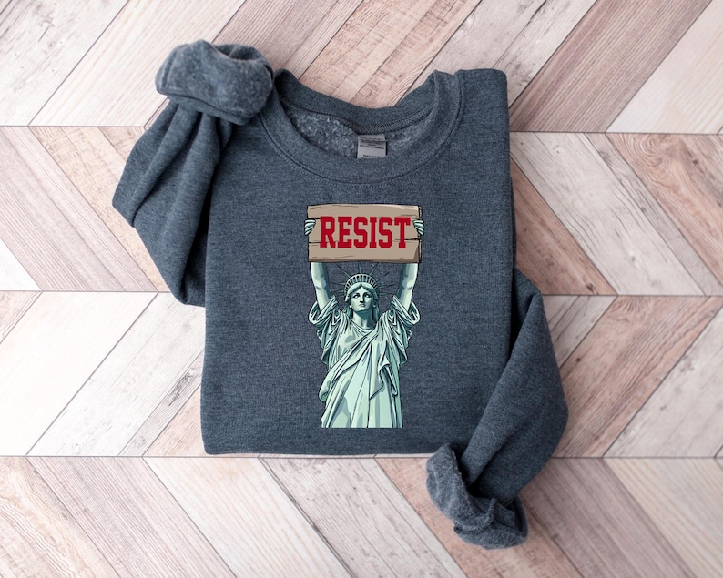 Op de afbeelding: Een donkergrijs sweatshirt met een afbeelding van het Vrijheidsbeeld dat een bord vasthoudt met de tekst "RESIST".