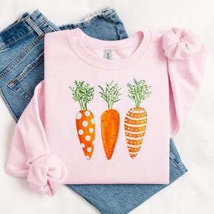 Puede incluir: Una sudadera rosa con un gr&aacute;fico de tres zanahorias naranjas con lunares blancos y rayas. Las zanahorias tienen hojas verdes.