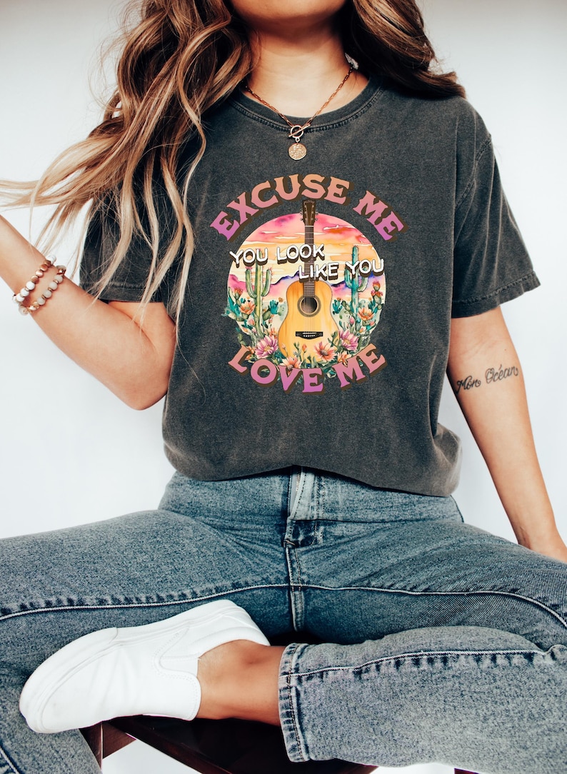 May include: A dark gray t-shirt with a graphic print of a sunset, cactus, and an acoustic guitar. The text on the shirt reads "Excuse Me You Look Like You Love Me".